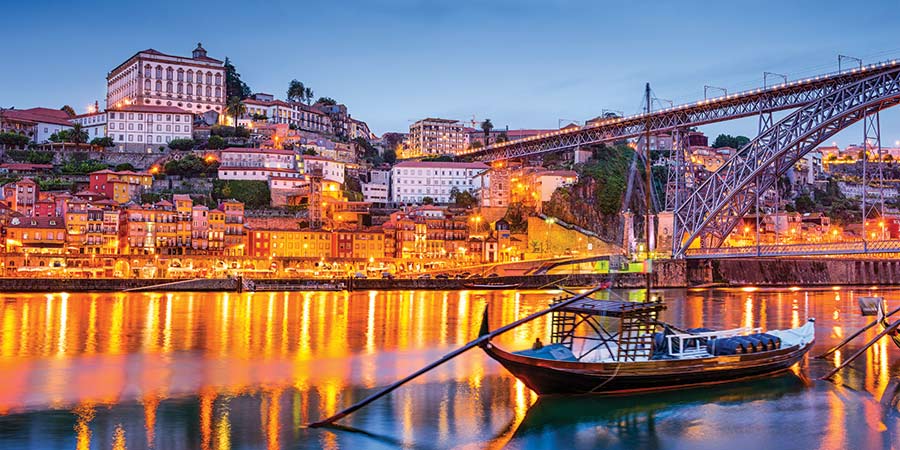 Porto is illuminated is warm orange light at night, a small river boat sits on the river and the Luís bridge can be seen. 